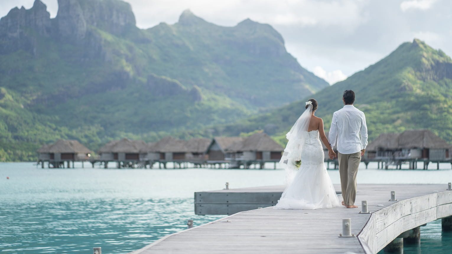 A couple walking hand in hand down an overwater jetty toward the mountains of Bora Bora, Four Seasons