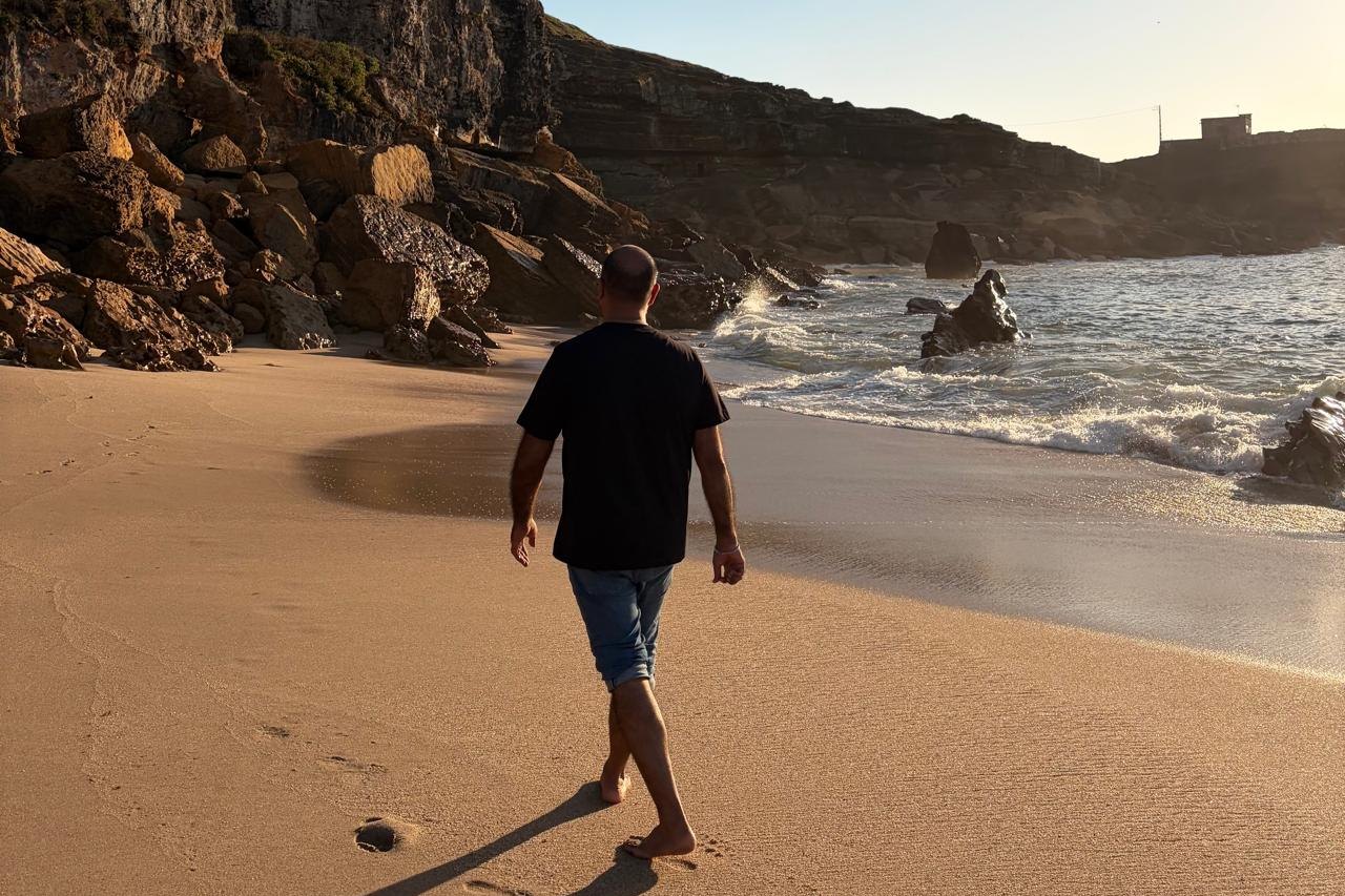 A man walking barefoot along a golden beach beneath tall cliffs as small waves roll in