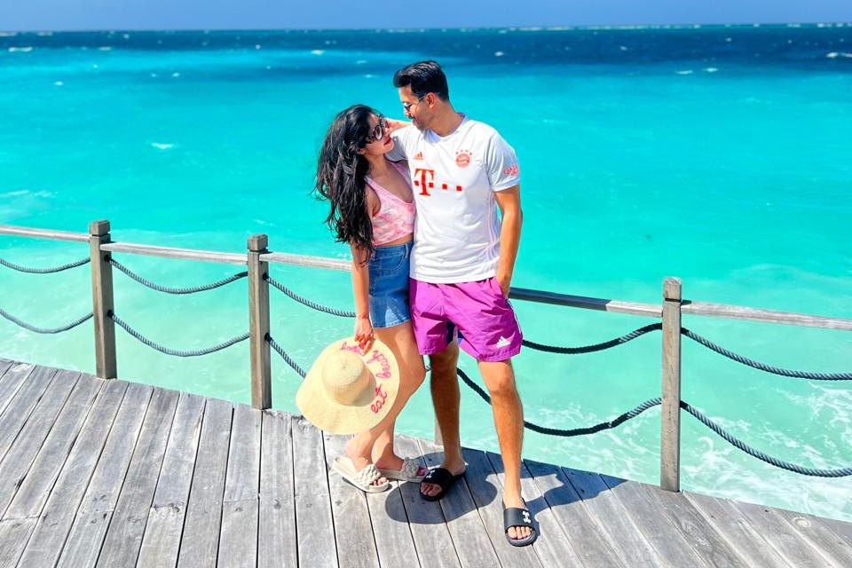 A couple standing close together on a wooden jetty above bright turquoise water, the woman holding a straw sun hat