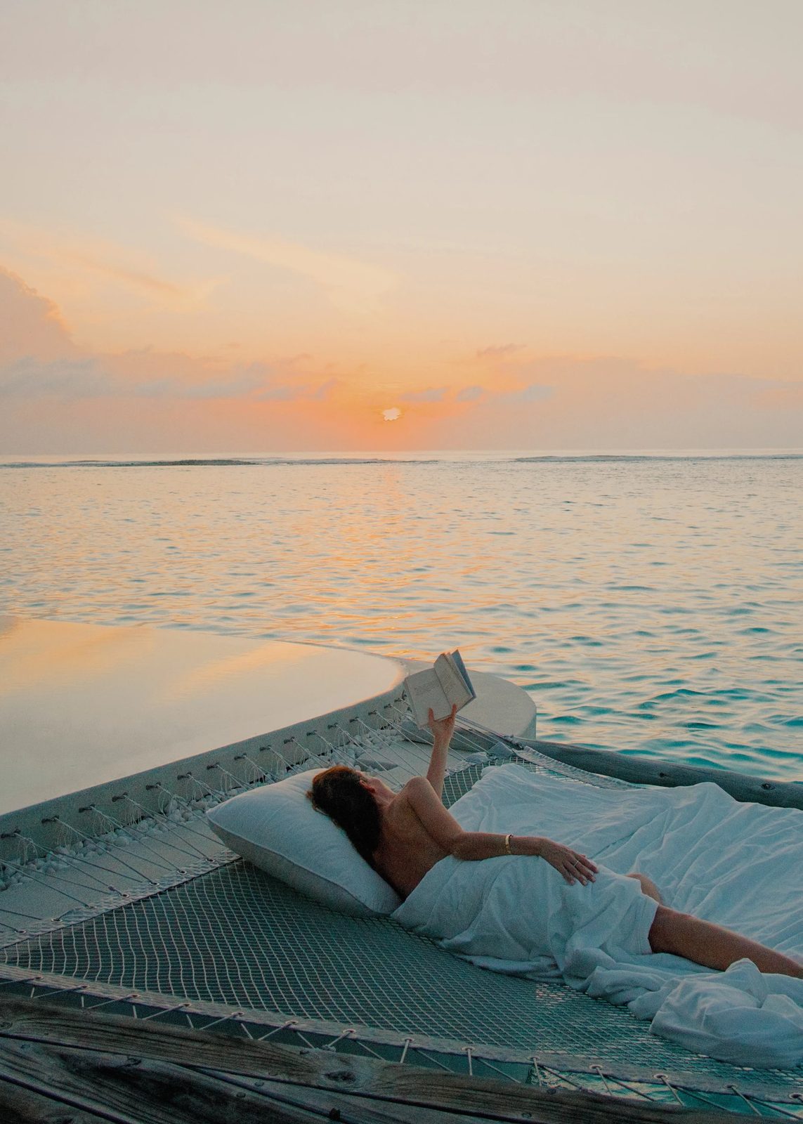 Woman reading at sunset on an overwater hammock above a turquoise Maldives lagoon
