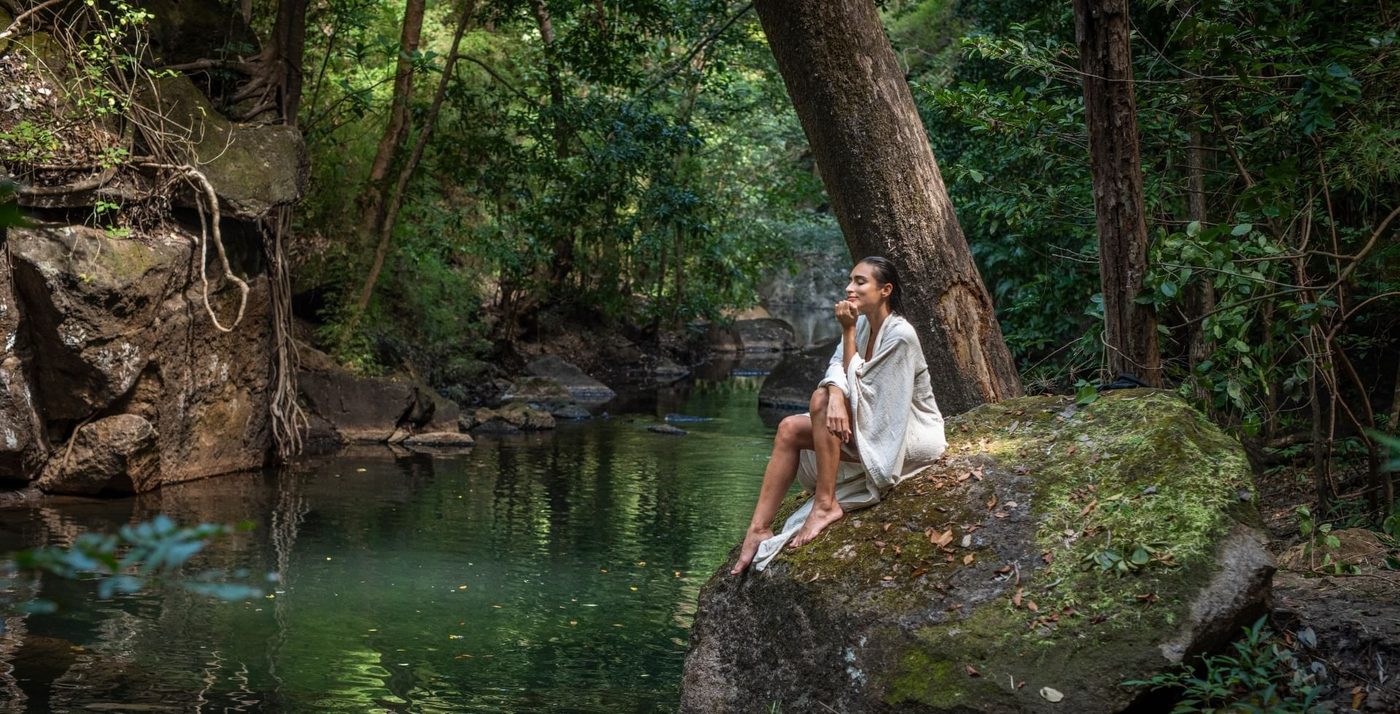 A guest wrapped in a soft robe, resting on a moss-covered rock beside the thermal river at Rio Perdido